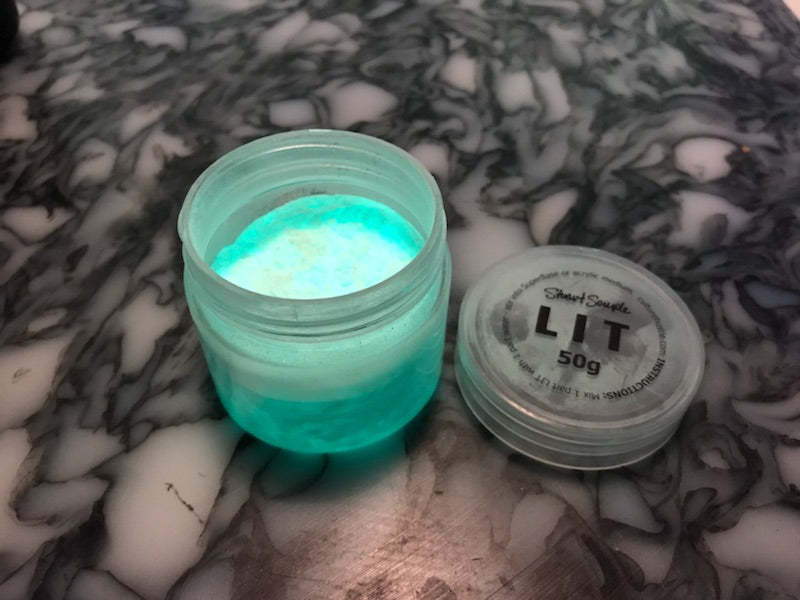 LIT glow pigment in a clear jar, showcasing turquoise powder inside, with a lid labeled "50g" by Stuart Semple.