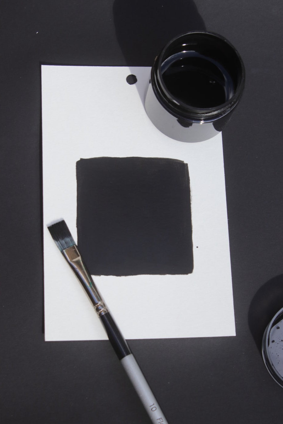 A black paint drawdown on white card beside an open jar and a flat brush